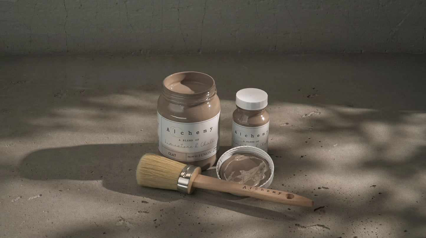 Botes Alchemy by Fusion Clay 500ml y 120ml con brocha redonda sobre superficie de cemento con sombras naturales, pintura a la tiza taupe gris cálido