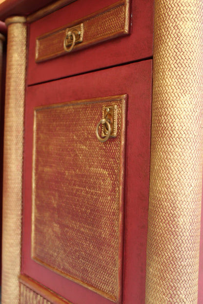 Detalle de la puerta y el cajón de una mesita vintage burdeos, con paneles decorativos dorados y tiradores de anilla.