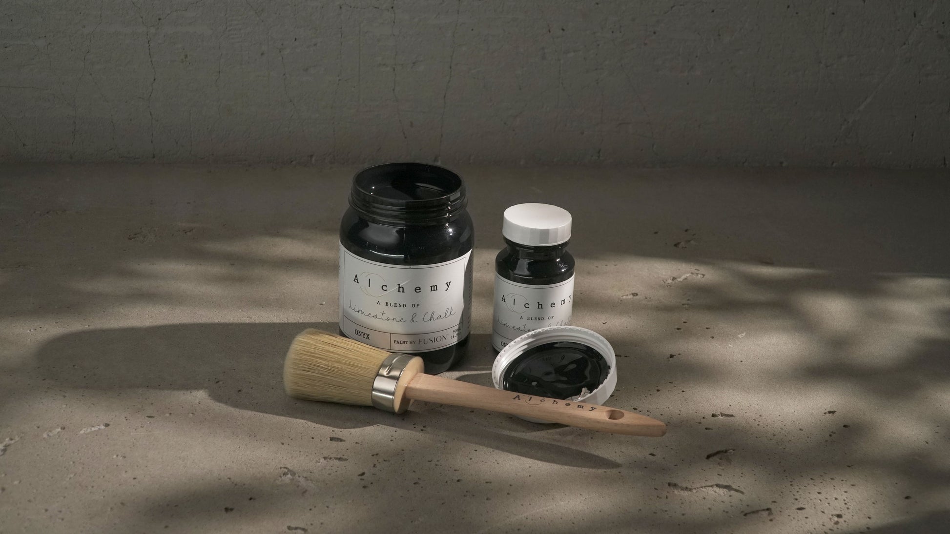 Botes Alchemy by Fusion Onyx 500ml y 120ml abiertos con brocha redonda sobre superficie de cemento con sombras de luz natural, pintura a la tiza negro puro