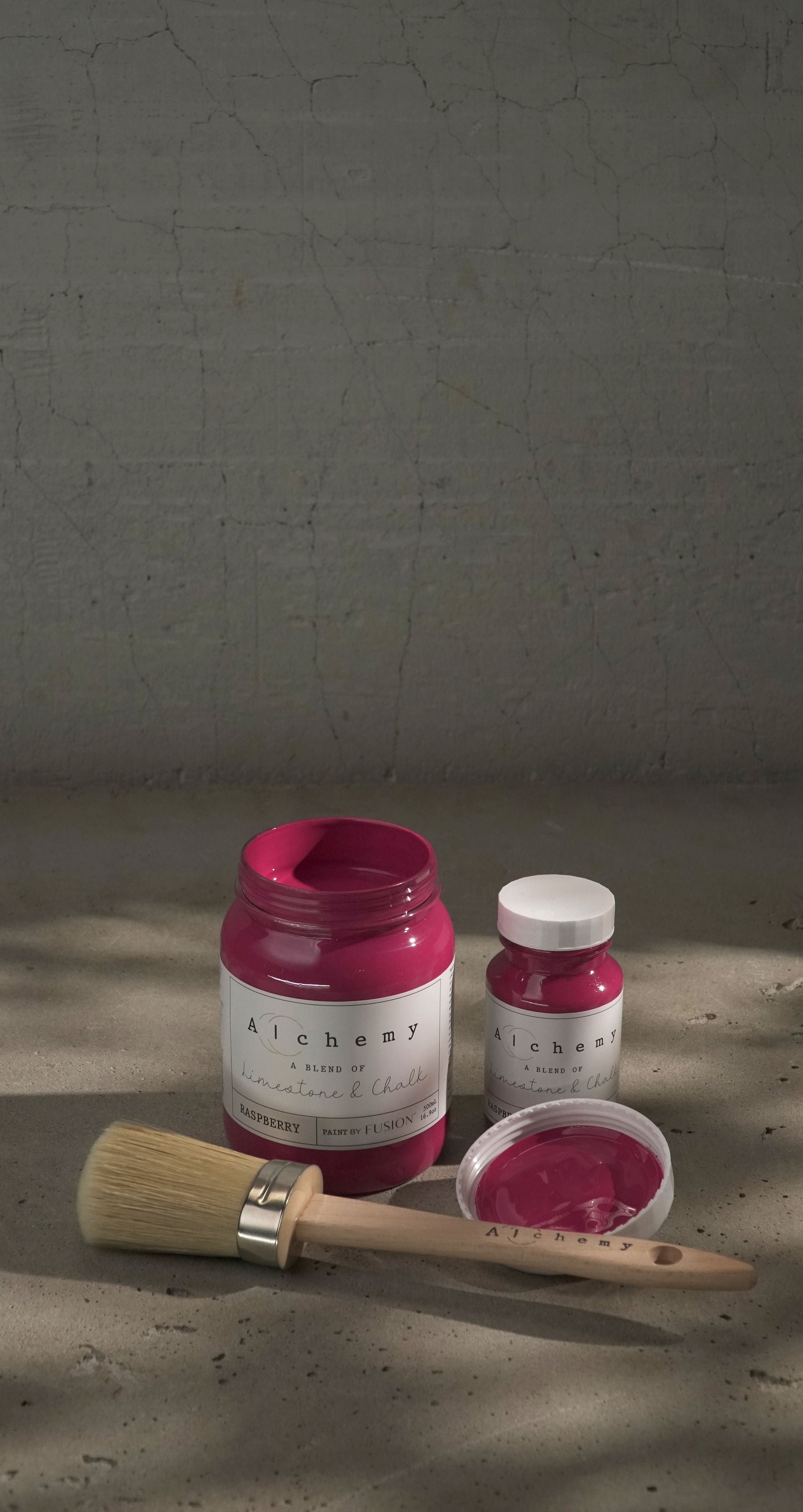 Botes Alchemy by Fusion Raspberry 500ml y 120ml abiertos con brocha sobre fondo de cemento texturizado con luz lateral, pintura a la tiza fucsia intenso limestone y chalk