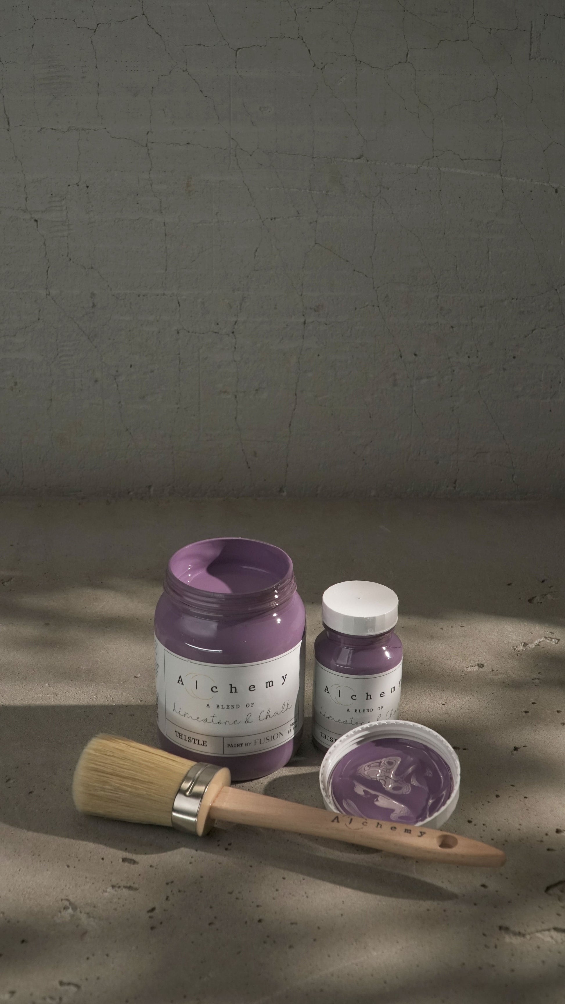 Botes Alchemy by Fusion Thistle 500ml y 120ml abiertos con brocha sobre fondo de cemento texturizado con luz lateral y sombras dramáticas, pintura a la tiza malva limestone y chalk