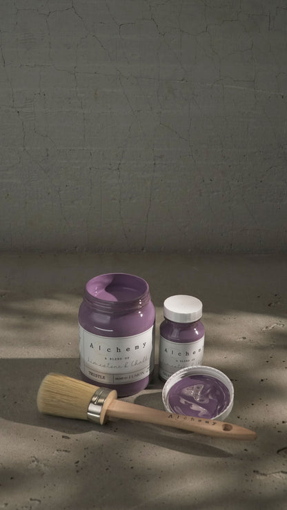 Botes Alchemy by Fusion Thistle 500ml y 120ml abiertos con brocha sobre fondo de cemento texturizado con luz lateral y sombras dramáticas, pintura a la tiza malva limestone y chalk
