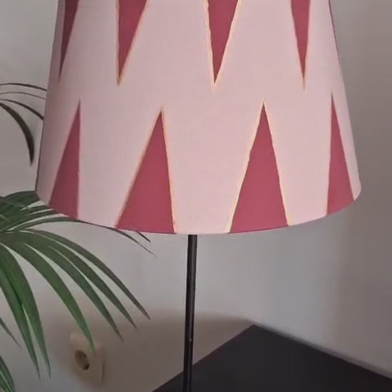 Pantalla de lámpara rosa empolvado con triángulos burdeos perfilados en dorado, presentada sobre una base negra junto a una planta.